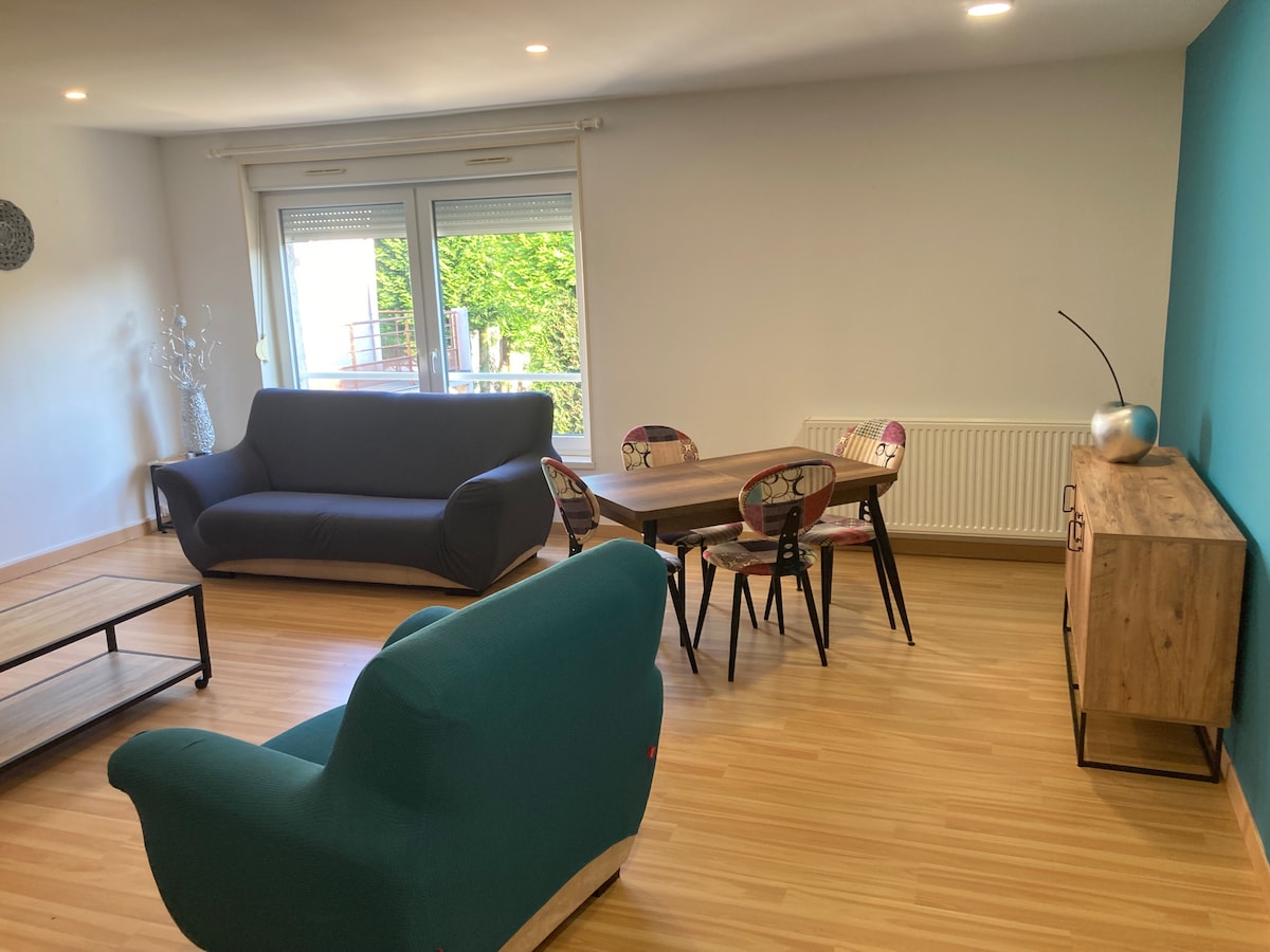A spacious living area is presented, featuring a dark sofa and a light blue armchair. A wooden dining table with six chairs sits centrally, accompanied by a sideboard. Large windows allow natural light to fill the space, with greenery visible outside.