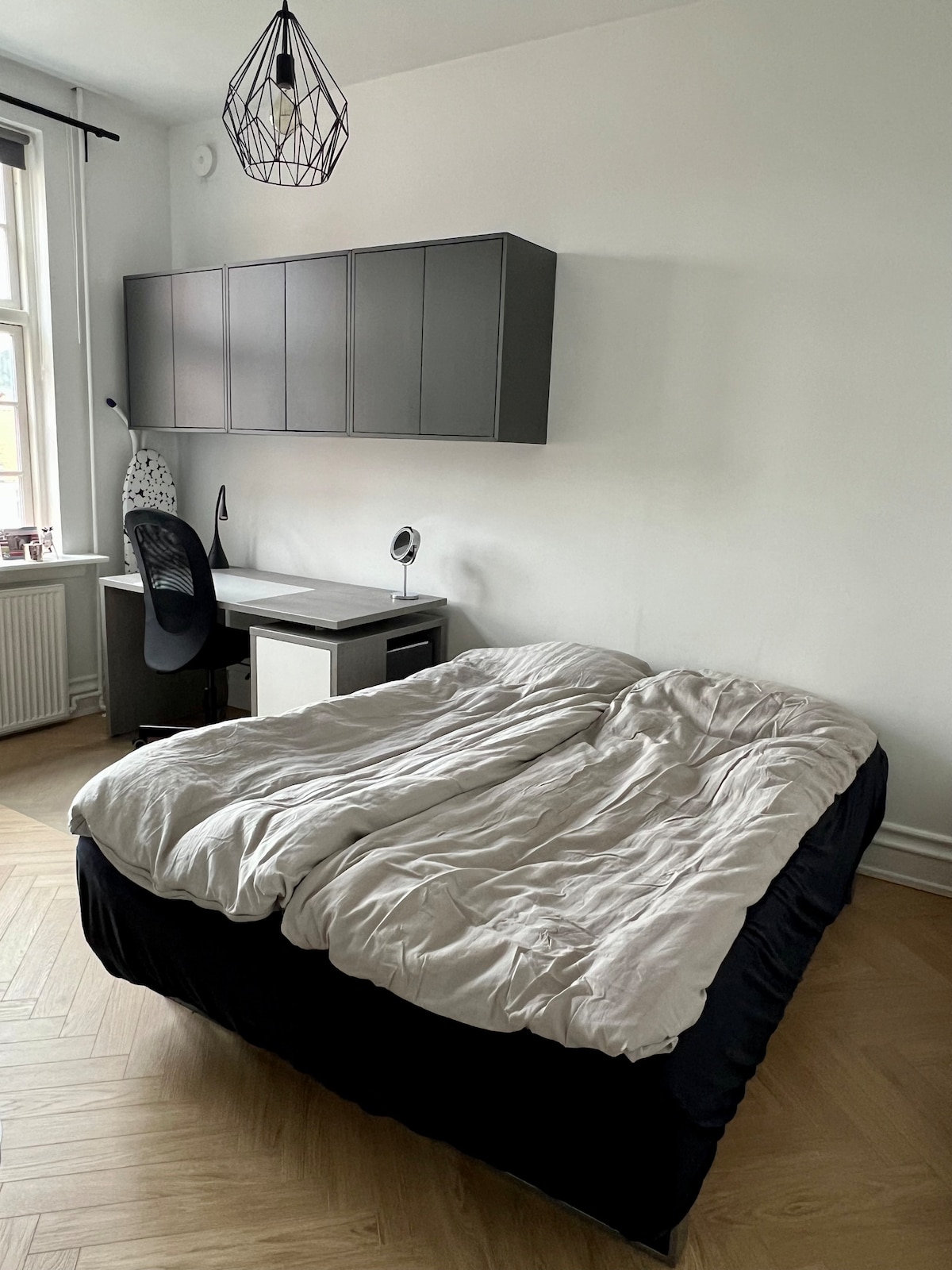 A spacious sleeping area features a large bed topped with a light-colored duvet. A modern desk and chair are positioned nearby, complemented by a stylish wall-mounted cabinet above. Natural light enters through a nearby window, illuminating the room's neutral tones.