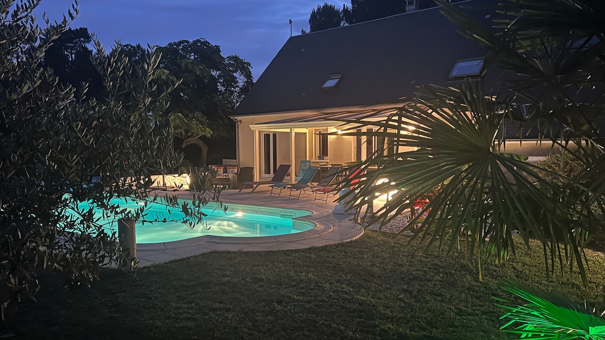 A spacious outdoor area features a heated swimming pool surrounded by lounge chairs, with soft lighting creating a calming atmosphere. Lush greenery, including palm trees, is visible around the pool, and the house showcases large windows illuminated by warm light in the evening.