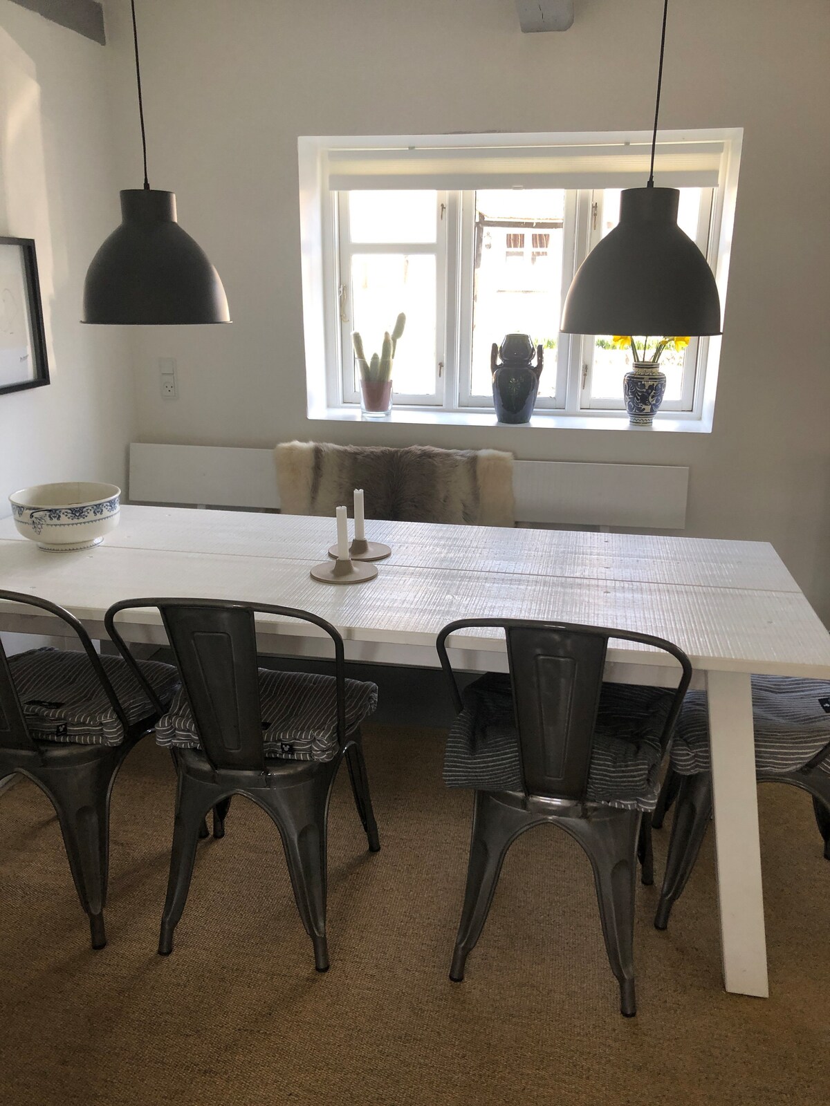 A clean dining area features a long white table surrounded by six metal chairs. Two black pendant lights hang above the table. A window with light drapes allows natural light to illuminate the space, highlighting decorative vases and a bowl on the table.