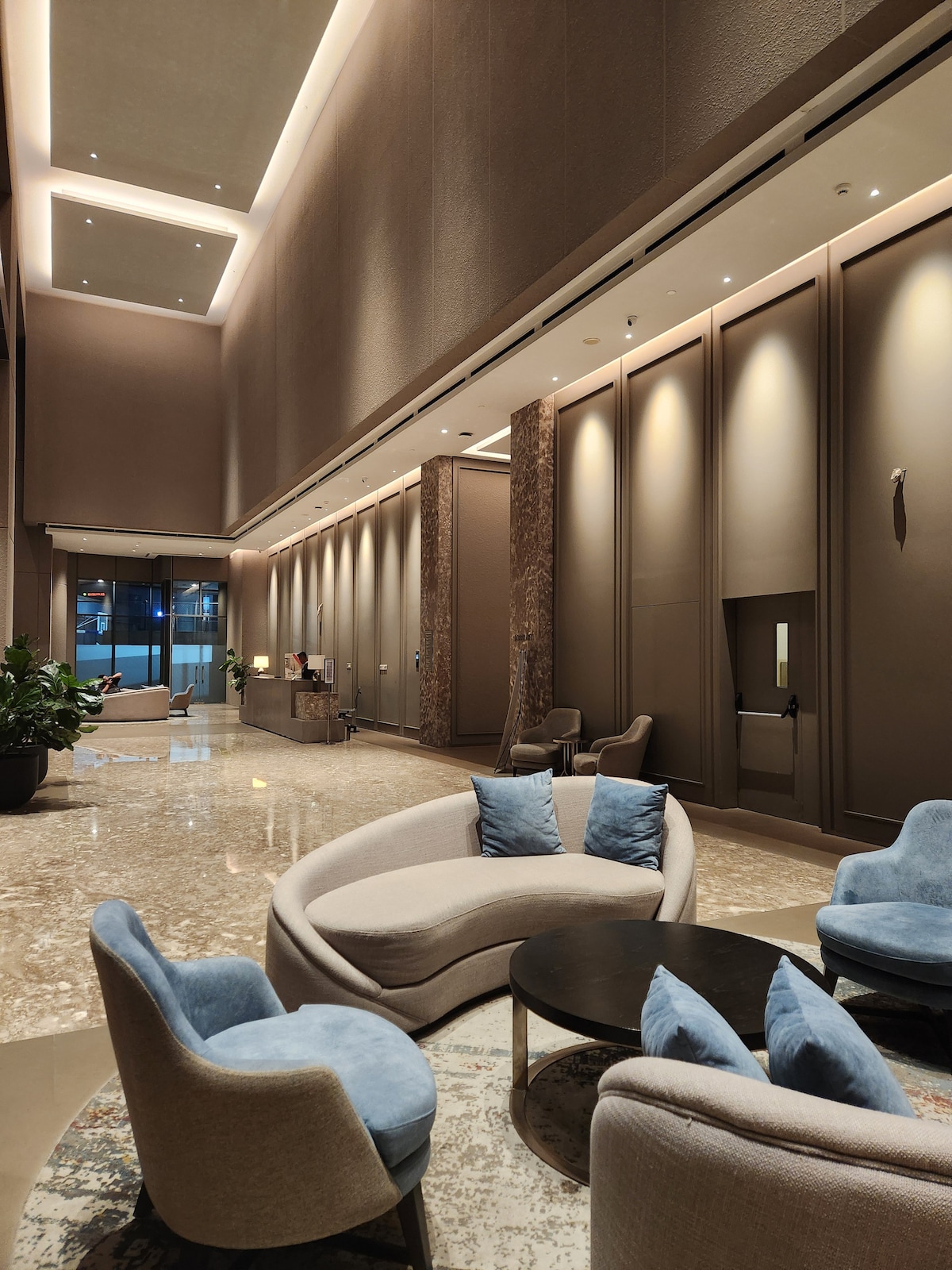 A modern lobby area is shown, featuring a smooth marble floor and high ceilings. Soft seating options in light gray and blue are arranged around a dark circular table. Soft lighting illuminates the walls, enhancing the contemporary design.
