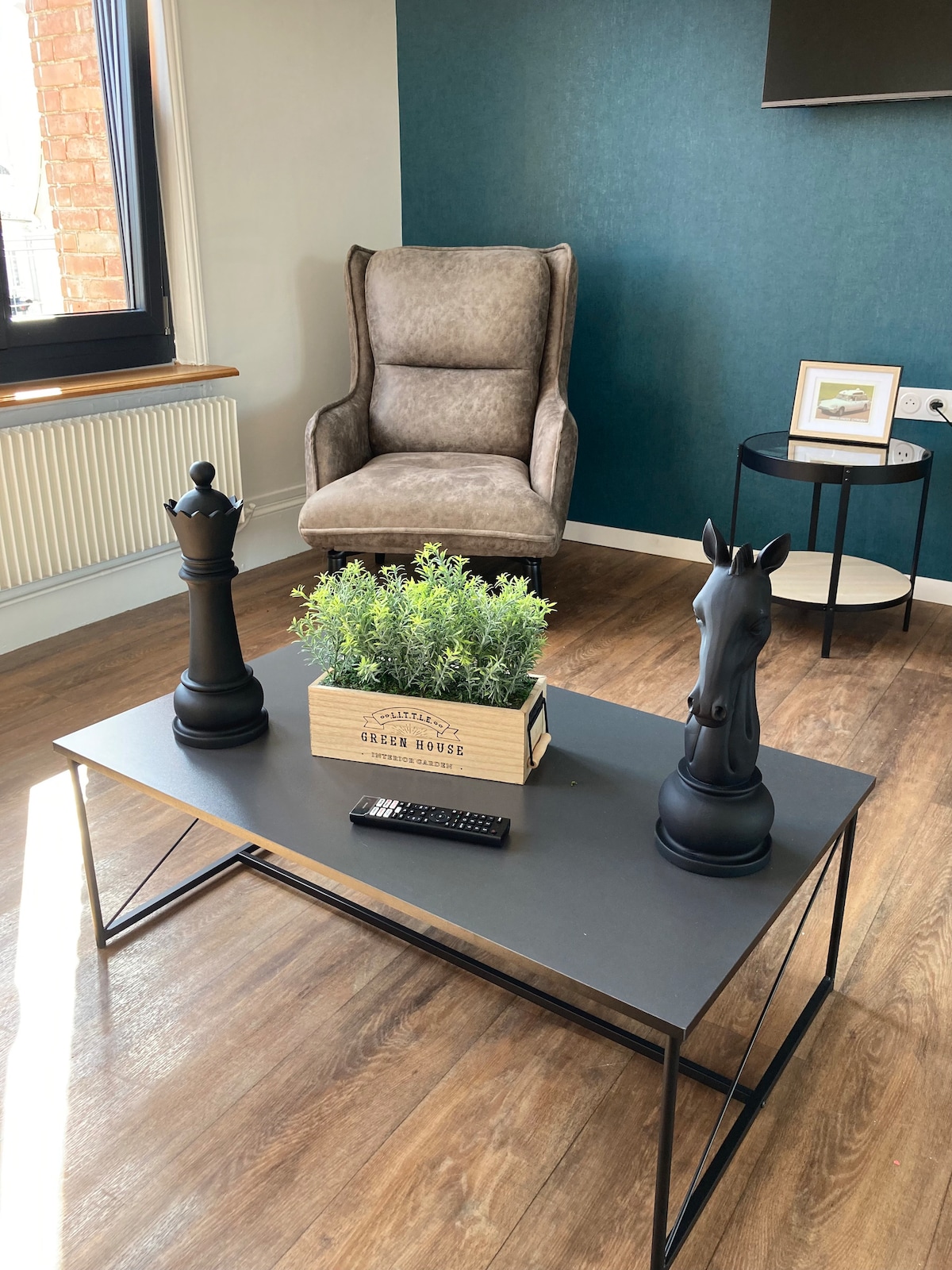 A modern sitting area features a sleek coffee table at the center, adorned with decorative plant arrangements and a remote control. Two artistic chess pieces, a king and a knight, flank the table, while a cozy armchair and a small side table complete the decor.