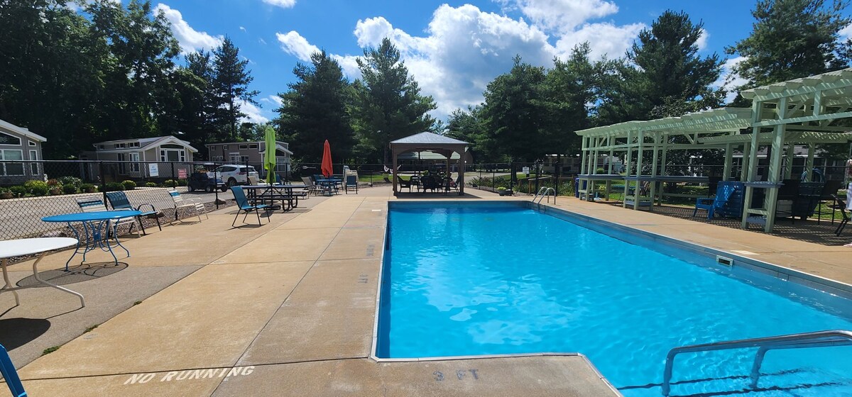 An outdoor swimming pool is set within a landscaped area featuring lounge chairs and shaded seating. A gazebo offers a communal gathering space. Sunlit trees surround the pool area, providing a serene environment for relaxation.