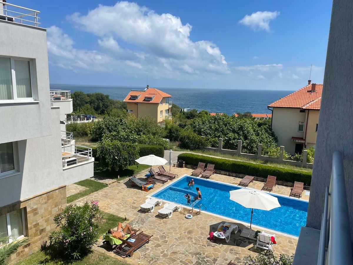 A view of the outdoor pool area is presented, featuring sun loungers arranged around the clear blue water. Umbrellas provide shade nearby. In the background, green shrubbery and distant buildings are visible under a partly cloudy sky, with the sea visible beyond.