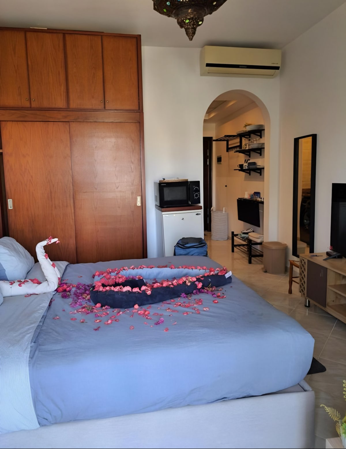 A spacious studio bedroom features a neatly made bed adorned with delicate flower petals. A modern television is positioned on a light wood cabinet, while a mirror reflects the room's natural light. A kitchenette and additional amenities are visible in the background.