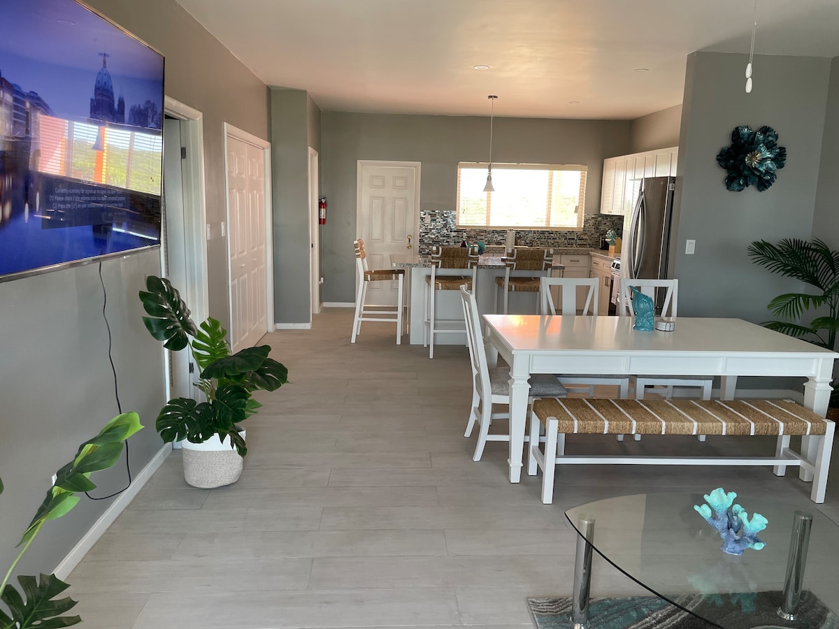 An open-concept living area is displayed, featuring a white dining table with wooden bench seating and a large television mounted on the wall. A modern kitchen is visible in the background, with light cabinetry and a tile backsplash. Greenery adds a touch of nature.