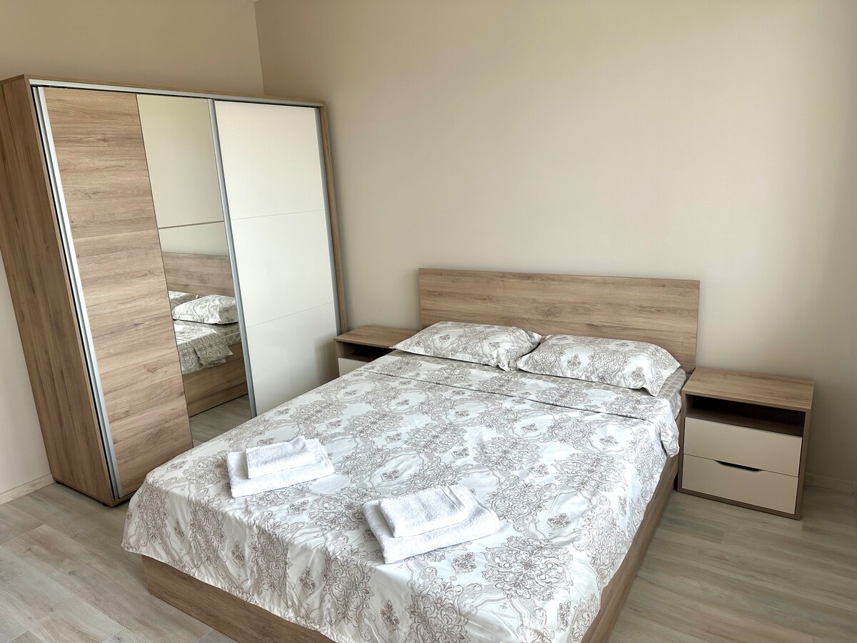A spacious bedroom features a large bed with a patterned duvet and neatly arranged white towels. A mirror-fronted wardrobe stands beside the bed, complemented by matching bedside tables. Soft natural light fills the room, highlighting the neutral color palette and clean lines of the furnishings.