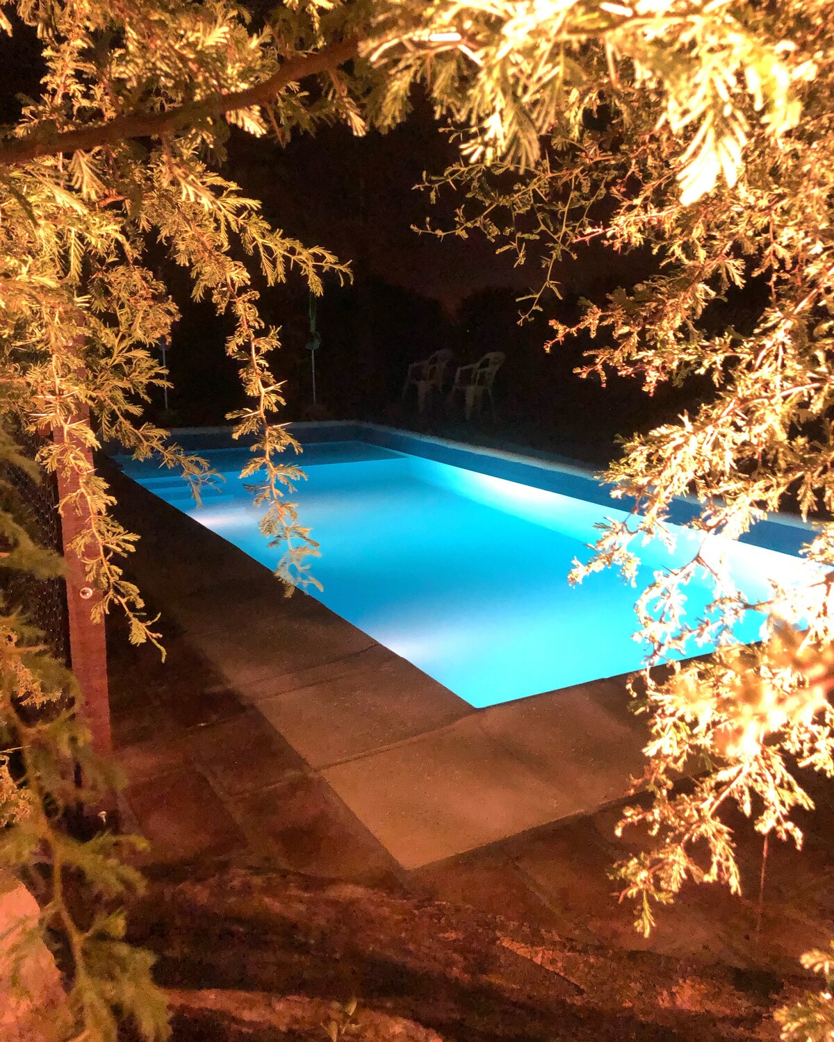 A tranquil swimming pool is illuminated with a soft blue glow under the night sky. Soft, green foliage frames the pool area, while two white chairs are positioned nearby, creating a serene space for relaxation.