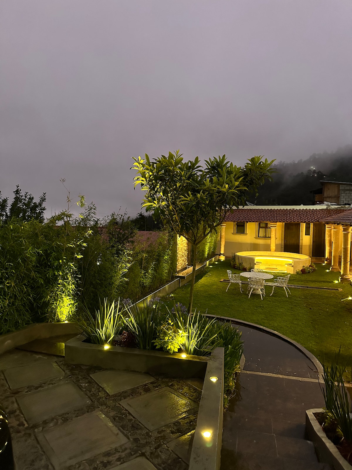 A serene garden area is illuminated softly, featuring a circular seating arrangement and a small dining table surrounded by greenery. Soft ambient lights highlight the garden's pathways, creating a relaxing outdoor space amidst the natural landscape.