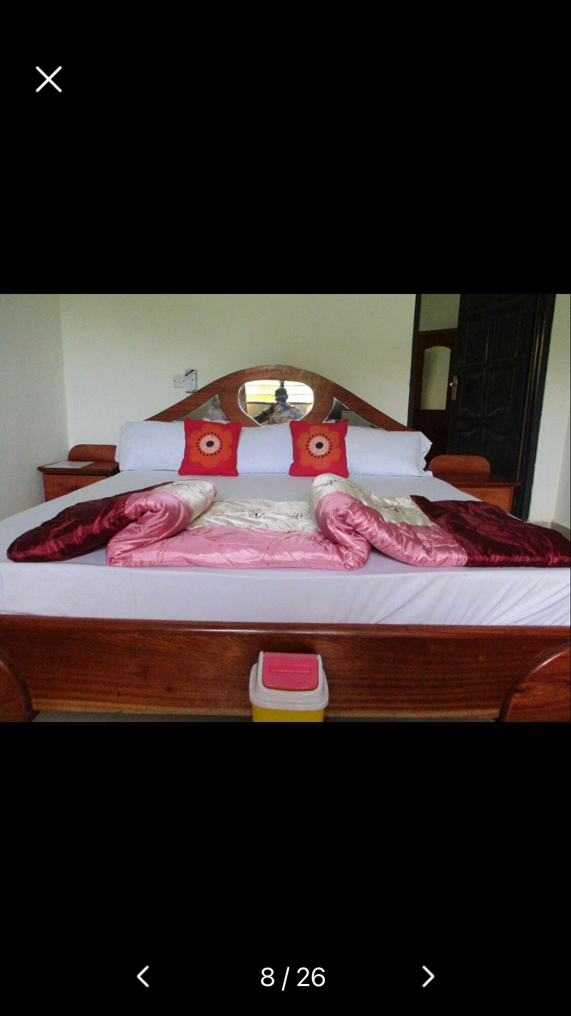 A spacious bed is centrally positioned in the room, adorned with decorative pillows featuring circular designs. A plush blanket in warm tones is neatly arranged across the bed, while wooden furniture adds to the overall aesthetic. Natural light filters in from an unseen window.