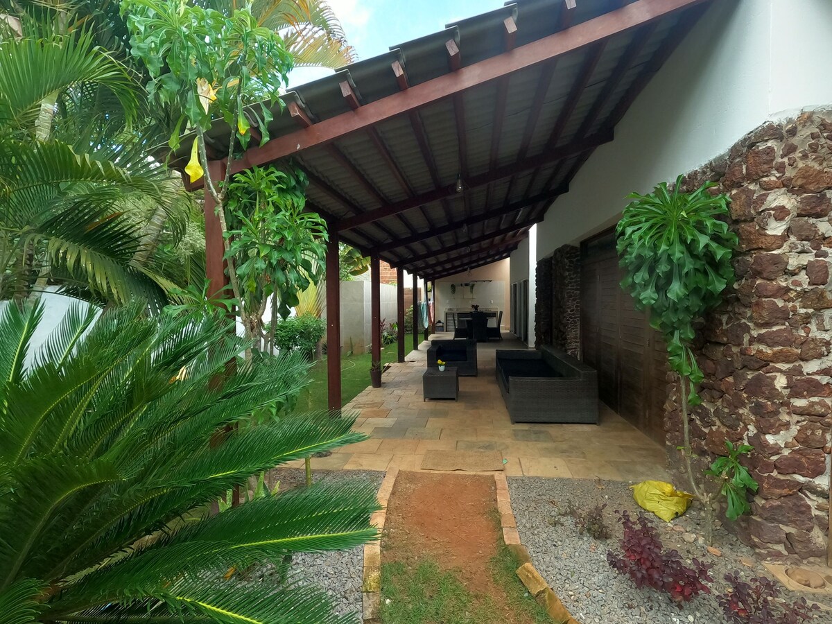 An outdoor area is shown, featuring a covered patio with comfortable seating arrangements. Lush greenery surrounds the space, enhancing the natural ambiance. A stone pathway leads through a landscaped yard, promoting a serene and inviting atmosphere for relaxation.