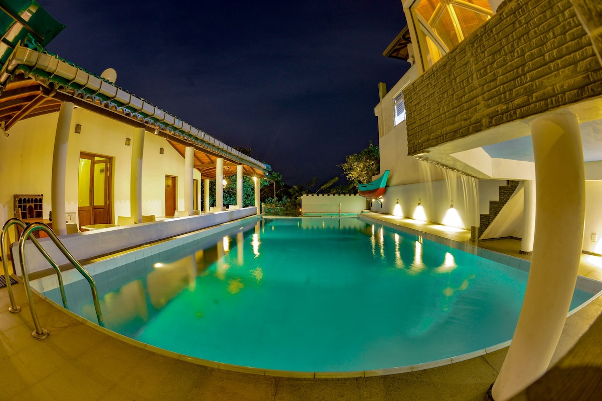 A serene pool area is illuminated softly at night, with inviting turquoise water reflecting surrounding lights. The space features simple elegant architecture, including large windows and lush greenery visible in the background, providing a tranquil atmosphere.