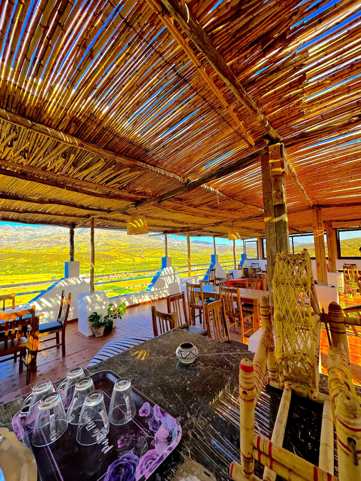An open dining area features bamboo roofing and is surrounded by a natural setting. Wooden tables and chairs are arranged across the space, with a view of expansive greenery visible through large openings. Glassware is neatly placed on a dark tabletop.