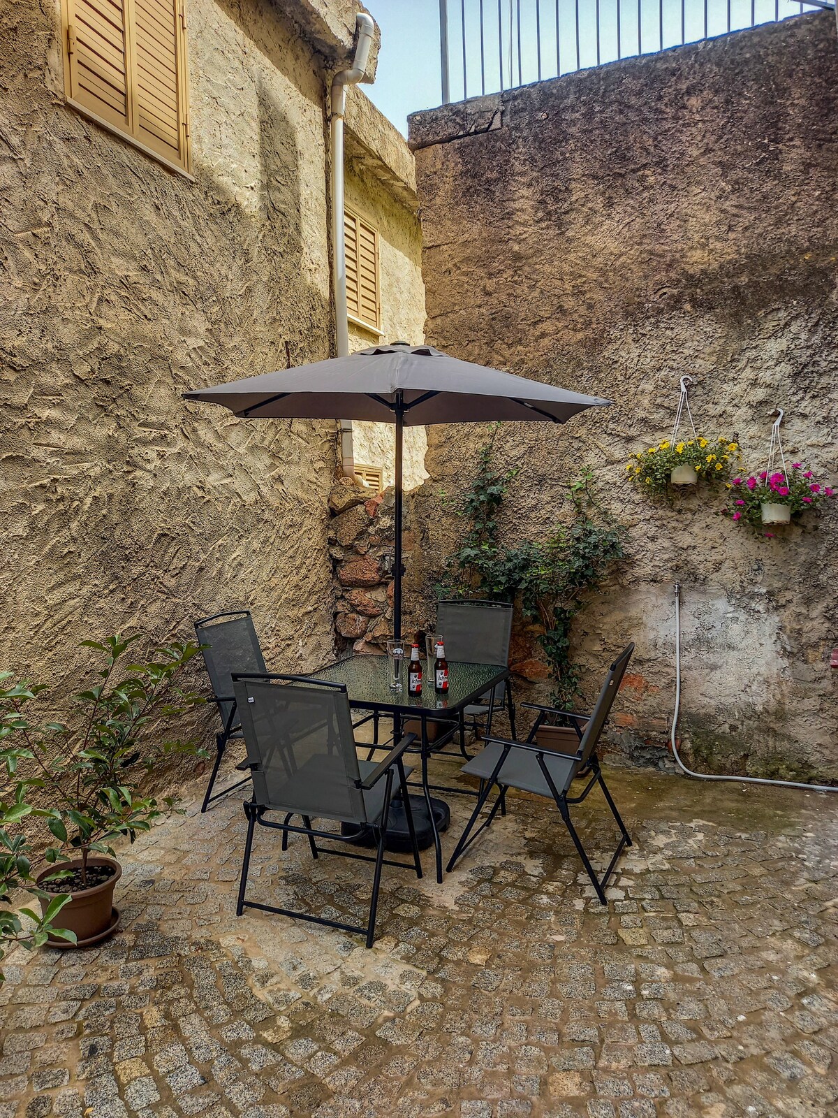 An outdoor area features a glass-top table surrounded by four black metal chairs, shaded by a large umbrella. The cobblestone floor creates a rustic charm, while walls adorned with climbing plants and hanging flower pots add a touch of greenery.