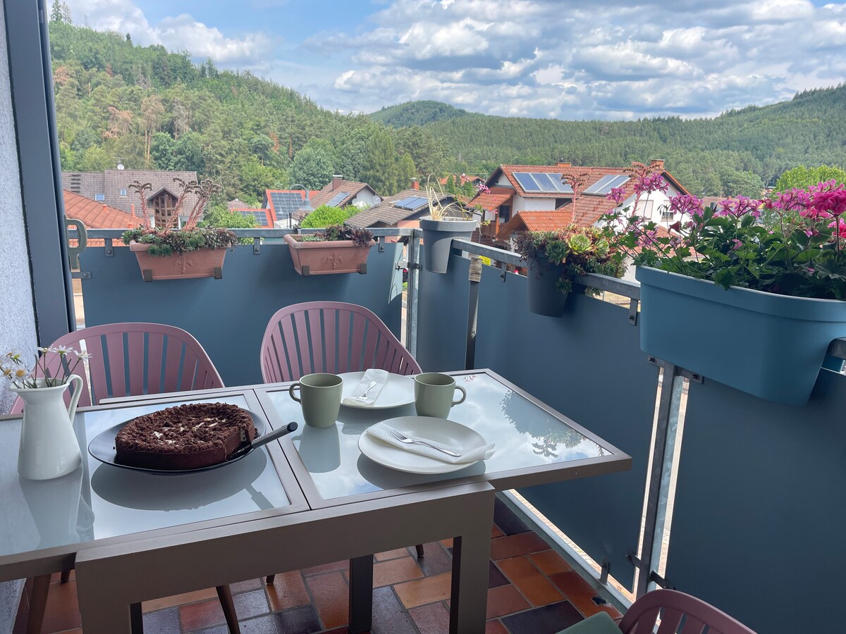An outdoor balcony is showcased with pastel chairs and a table set for two, featuring a dessert on a plate. Flower pots adorn the railing, and a view of lush green hills and homes in the distance can be seen under a partly cloudy sky.