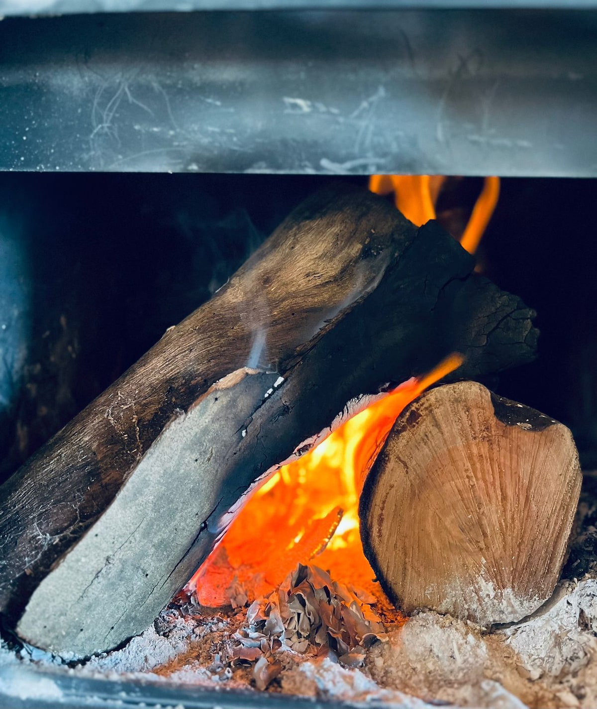 A cozy fireplace is shown with logs burning steadily. Flames flicker warmly against a backdrop of glowing embers and soft ashes, creating a serene atmosphere. The logs display natural textures and colors, enhancing the inviting feel of the space.