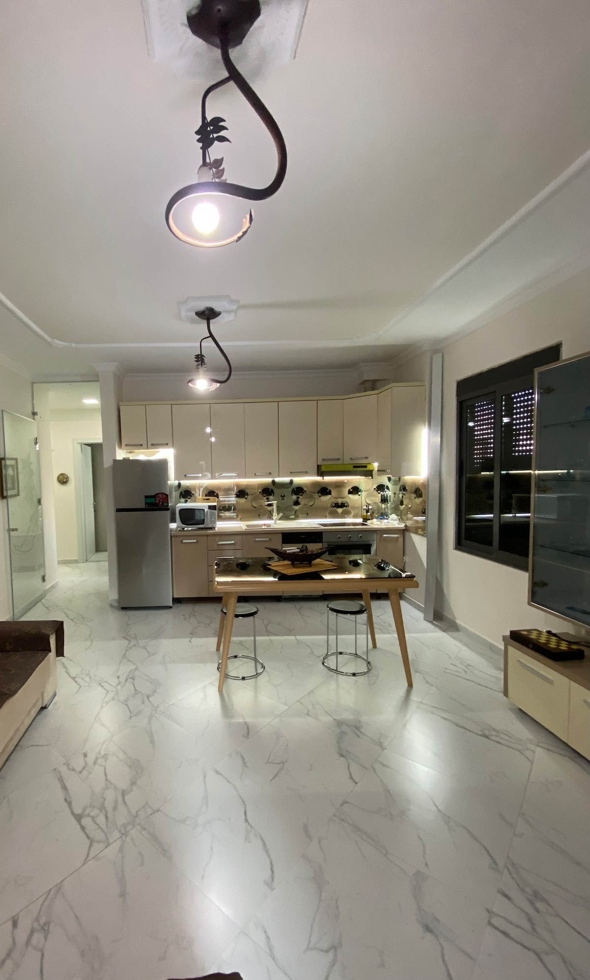 A modern kitchen featuring light-colored cabinetry and a marble floor. A small wooden table with two stools occupies the center. Stainless steel appliances and a well-lit counter add functionality, while natural light filters through a window with dark accents.