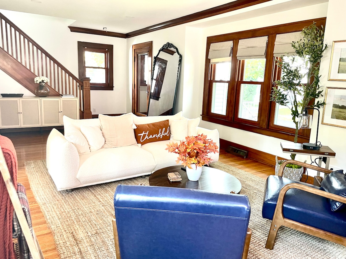 A cozy living room is depicted with a soft white sofa adorned with a decorative pillow. A round coffee table sits in front, featuring a small plant and decorative items. Large windows framed in rich wood allow natural light to illuminate the space, enhancing the warm atmosphere.