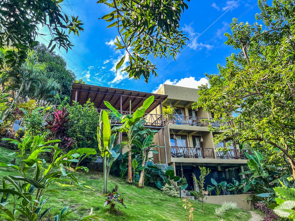 The exterior of a three-story building is surrounded by lush greenery, showcasing a variety of tropical plants and trees. The structure features multiple balconies with wooden railings, providing a connection to the natural landscape. Bright blue skies create a vibrant backdrop.