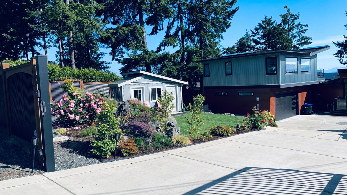 A landscaped outdoor area features a mix of greenery and flowering plants, along with a small blue garden shed. A gravel pathway leads from a brown wooden gate, with a modern house visible in the background, surrounded by tall trees and sunlight.