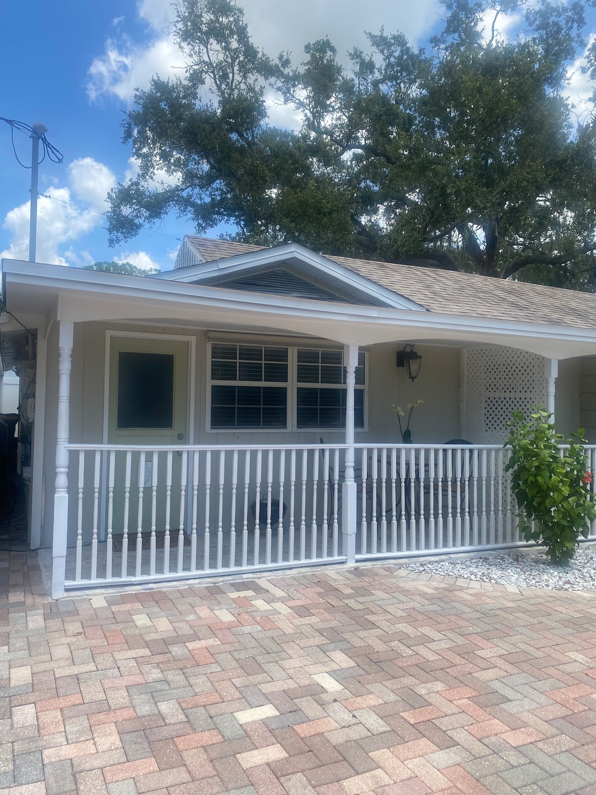 A charming exterior view of a single-story house is presented, showcasing a covered porch with white railings and an open door. Pavers are laid in a neat pattern on the pathway, while lush greenery is noticeable near the entrance, with spacious trees providing dappled sunlight.