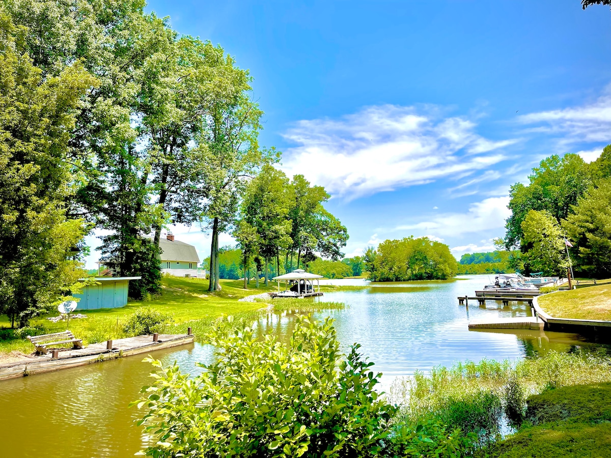 A scenic view of a calm lake surrounded by lush trees and greenery is presented. The vibrant blue sky reflects on the water, while a boat dock and gazebo are visible on the shore, enhancing the tranquil lakeside atmosphere.
