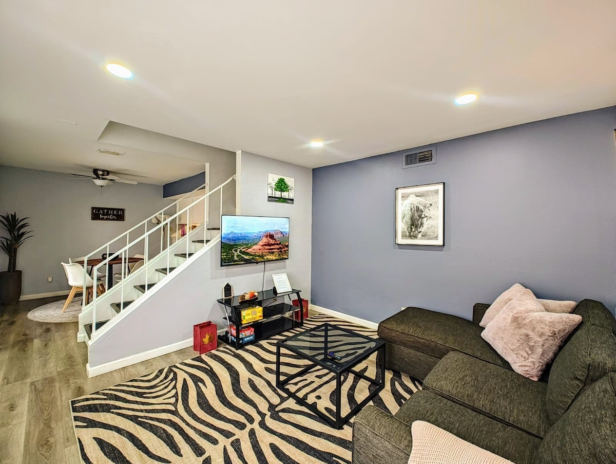 A cozy living area features a dark sectional sofa complemented by neutral cushions. A coffee table sits on a striped rug, and a television displays scenic imagery. A staircase leads to the upper level, while a dining area is visible in the background.