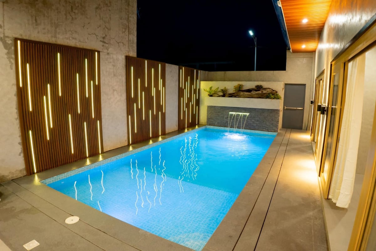 A modern swimming pool is illuminated under the night sky, surrounded by textured walls featuring vertical wooden accents. Soft lighting reflects off the water's surface, creating a serene ambiance. The pool area includes minimalist design elements that enhance a tranquil outdoor experience.
