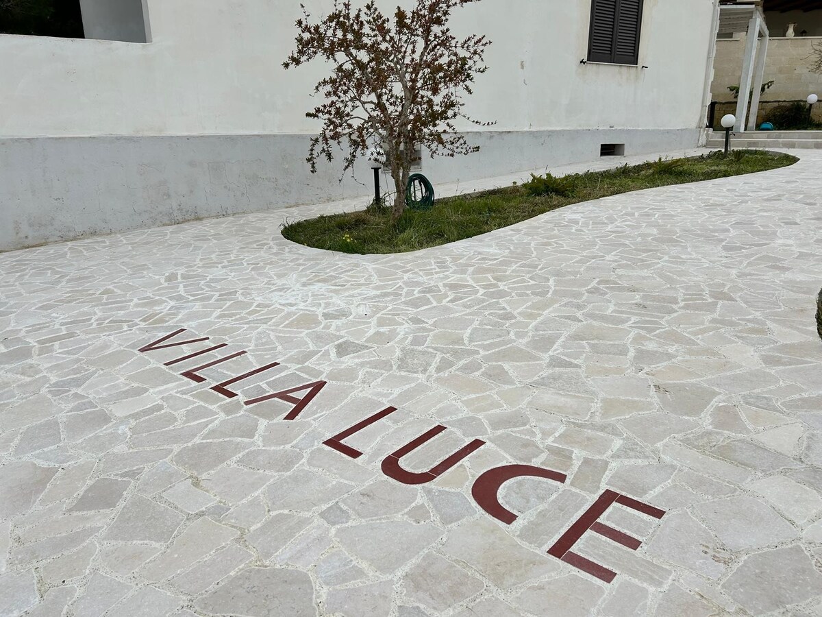 The pathway leading to the entrance of the property is made of light-colored stone, featuring the name 'VILLA LUCE' elegantly inscribed in red. The surrounding area includes green grass and a small decorative tree.