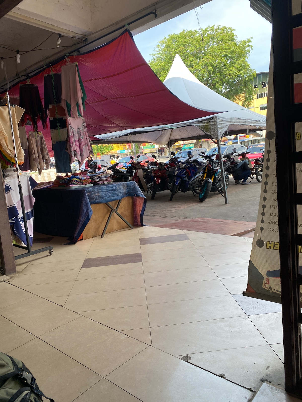 A vibrant market scene is visible through an entrance, featuring colorful fabric hanging from above. Beneath a large canopy, various motorcycles are parked. The tiled floor transitions from light to dark shades, leading to the bustling area outside filled with additional vendor stalls.