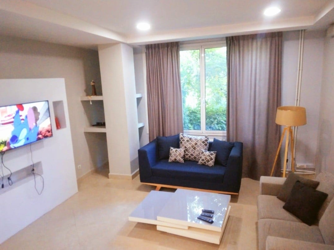 A modern living room features a navy blue couch adorned with decorative pillows facing a flat-screen TV mounted on a white wall. Natural light streams in through a large window, with partially folded curtains. A gray sofa complements the contemporary design, and a coffee table sits centrally.