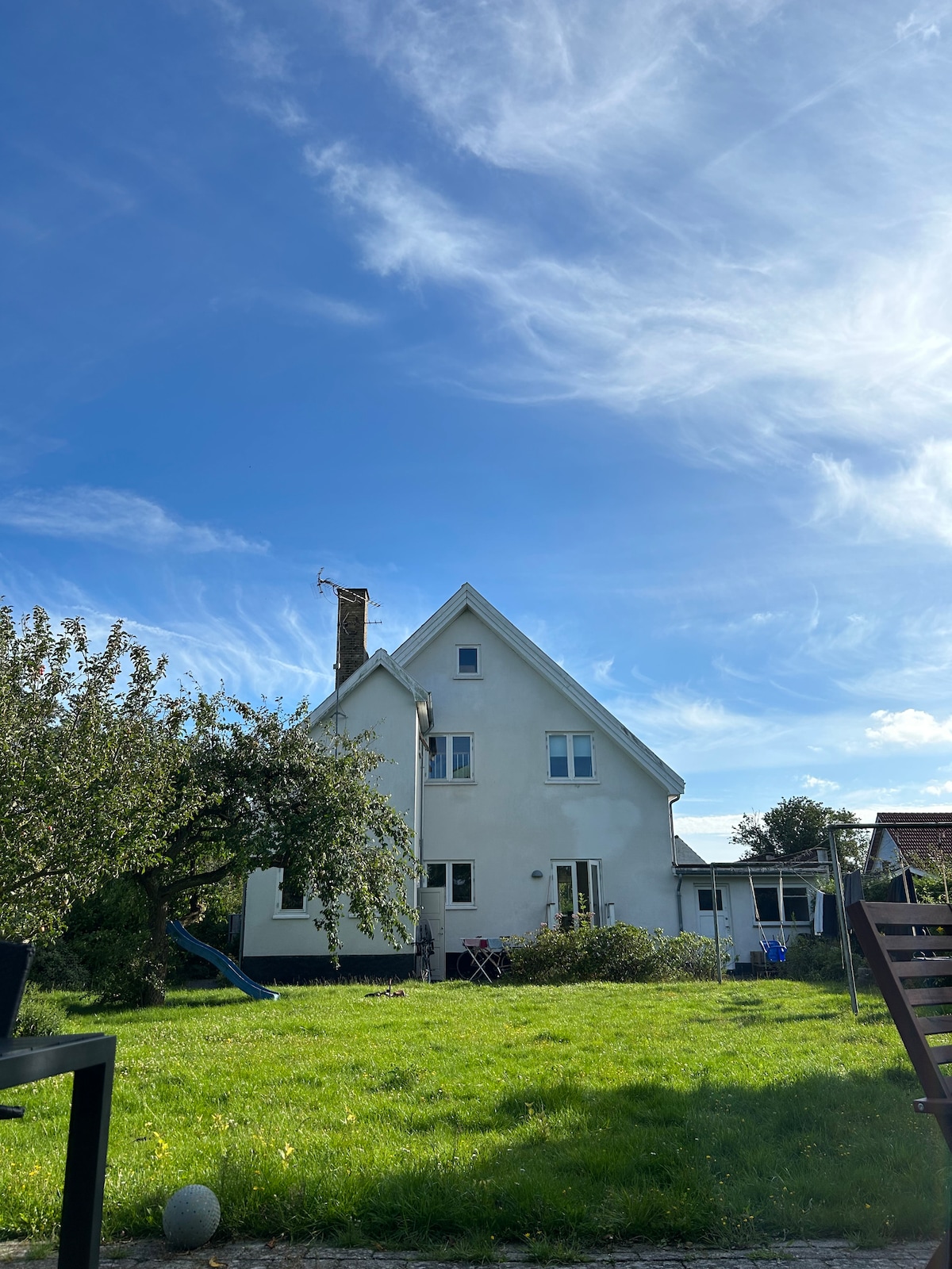 The house is presented from the garden, showcasing its white exterior against a bright blue sky. A grassy area is seen in the foreground, complemented by an apple tree and a wooden swing set. The structure features multiple windows, highlighting its inviting layout.