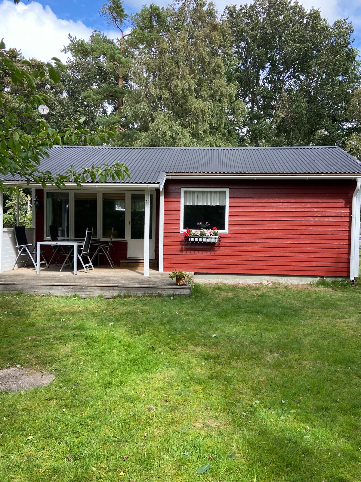 A small red and white summer cottage is set against a backdrop of greenery. A wooden deck leads to a patio with a table and chairs, while flower boxes with vibrant blooms add a charm to the exterior. The cottage features a sloped metal roof.