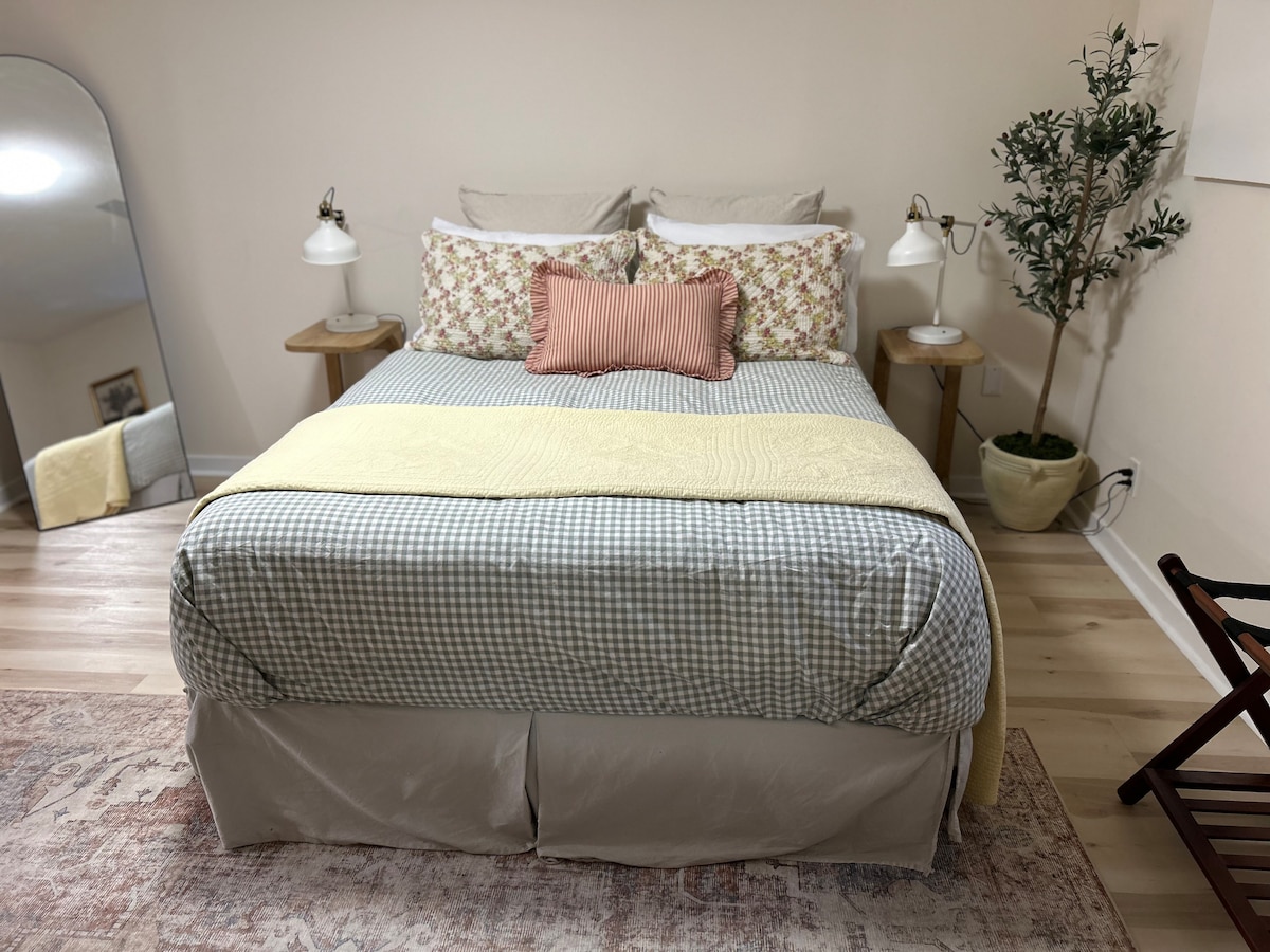 The cozy bedroom features a spacious bed with a blue and white checked blanket, adorned with decorative pillows. Two bedside lamps provide soft lighting. A tall mirror reflects the room, and a small potted plant adds a touch of greenery.