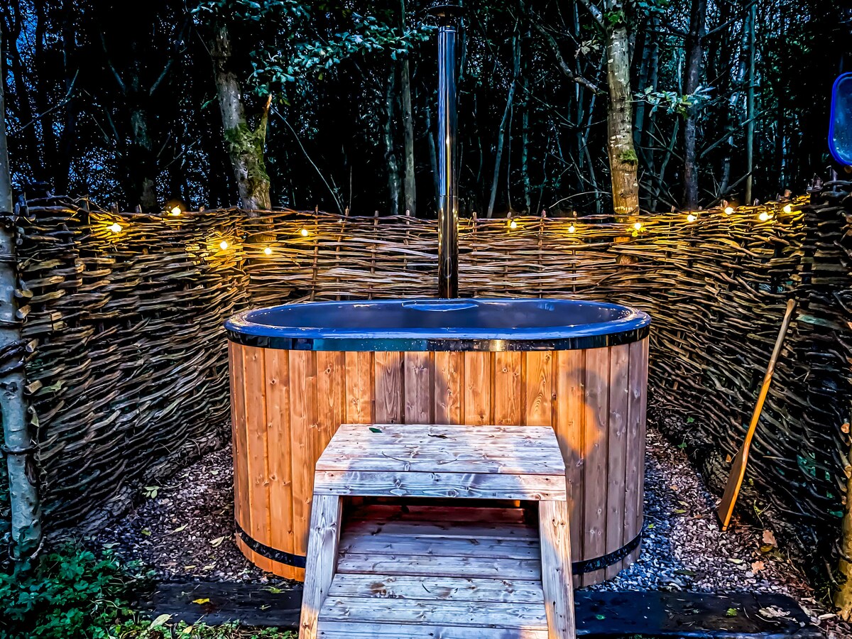 A log-fired hot tub bath is surrounded by a rustic woven fence and softly lit with string lights, creating a cozy atmosphere in a woodland setting. The tub features a wooden exterior and is positioned on a gravel surface, with a wooden step for easy access.