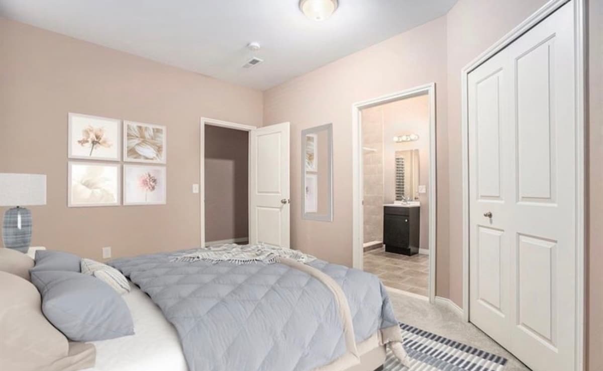 A bedroom with soft beige walls contains a spacious bed dressed in a gray quilt. Framed artwork decorates one wall, while light gently enters through a nearby door leading to a bathroom. Two doors are visible, providing access to other areas of the space.