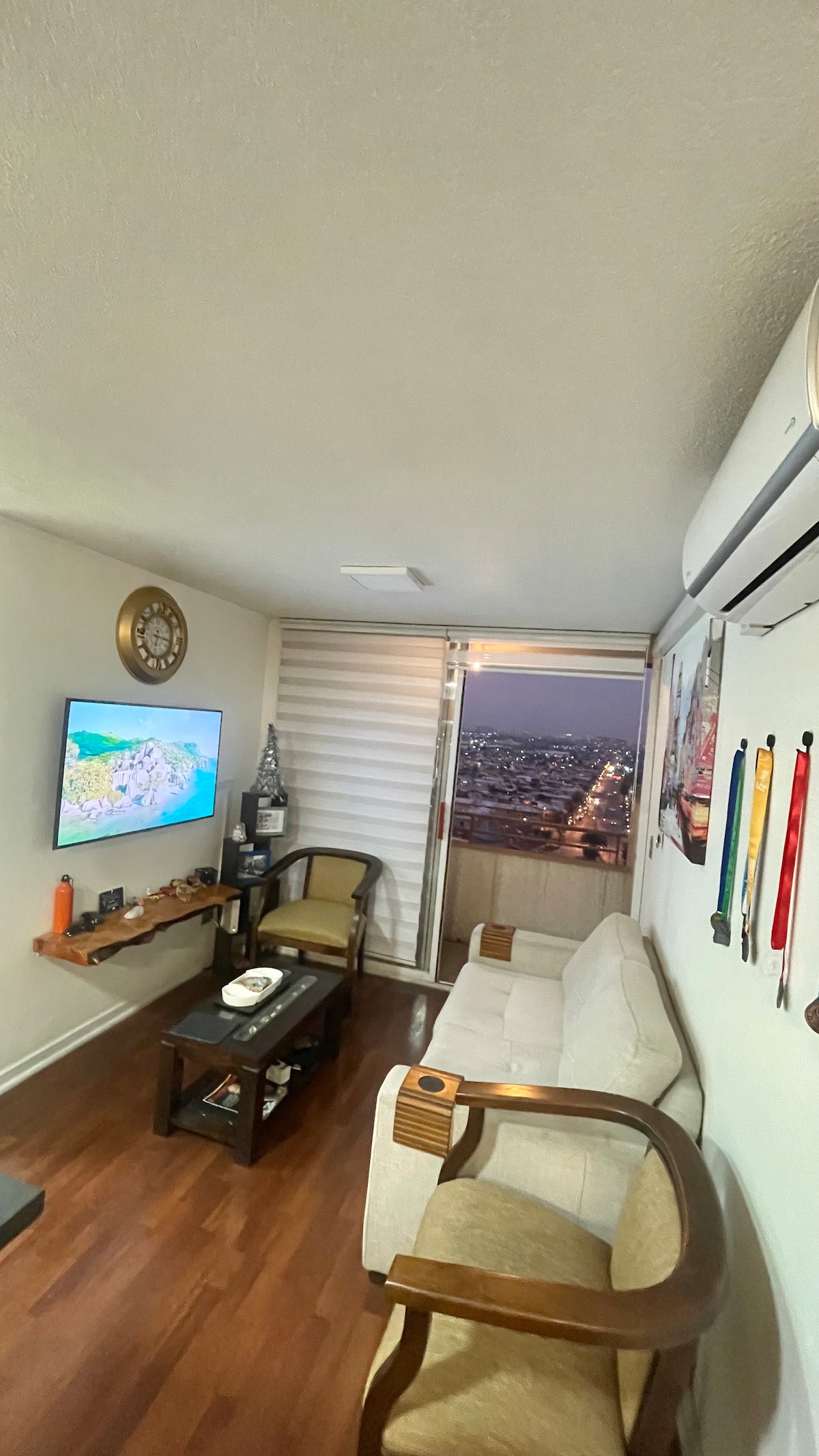 A cozy living area is presented featuring a neutral sofa and a wooden coffee table. A television is mounted on the wall, with a scenic view visible through the window covered by horizontal blinds. Decorative wall hangings and awards are displayed nearby.