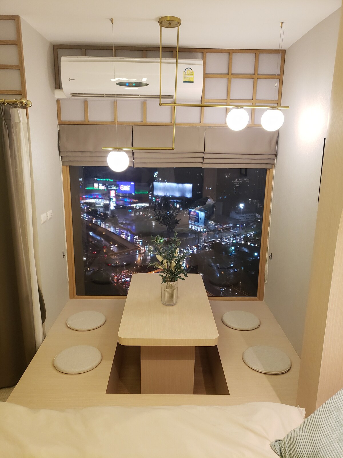 A dining area is positioned by a large window, offering a view of the city lights below. A round table is surrounded by four cushioned stools. A small vase with flowers sits at the center, while pendant lights provide soft illumination.