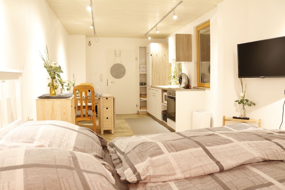 The interior features a modern, open layout with a cozy sleeping area, dining table, and kitchenette. Soft lighting enhances the neutral tones of the walls and furniture. A large window allows natural light to fill the space, while a television is mounted on one wall.