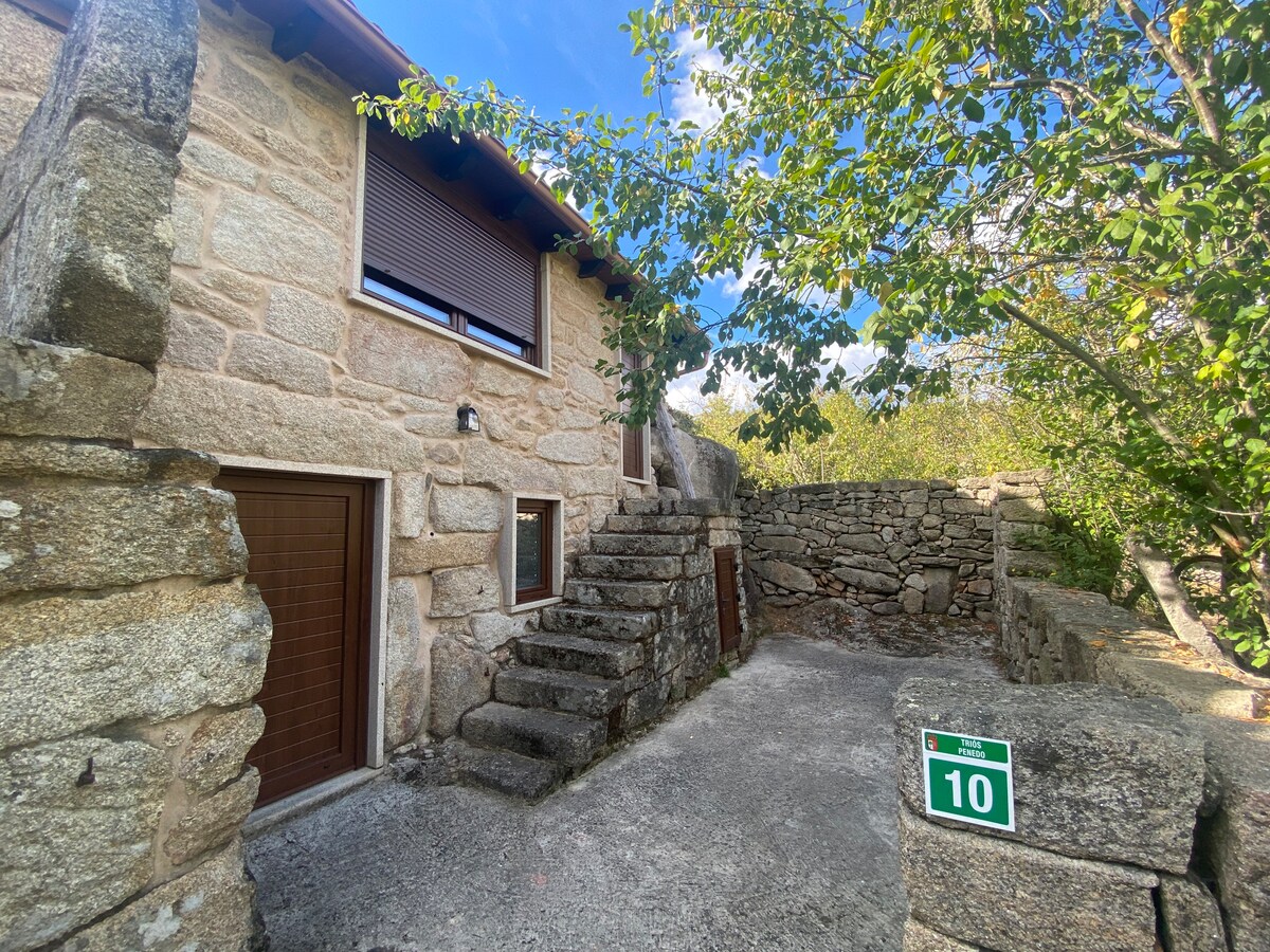 The image presents a charming stone house with wooden accents, featuring steps leading to an upper entrance. Natural surroundings include trees and carefully arranged stone walls, enhancing the rural appeal. A house number sign is visible, contributing to a welcoming entryway.