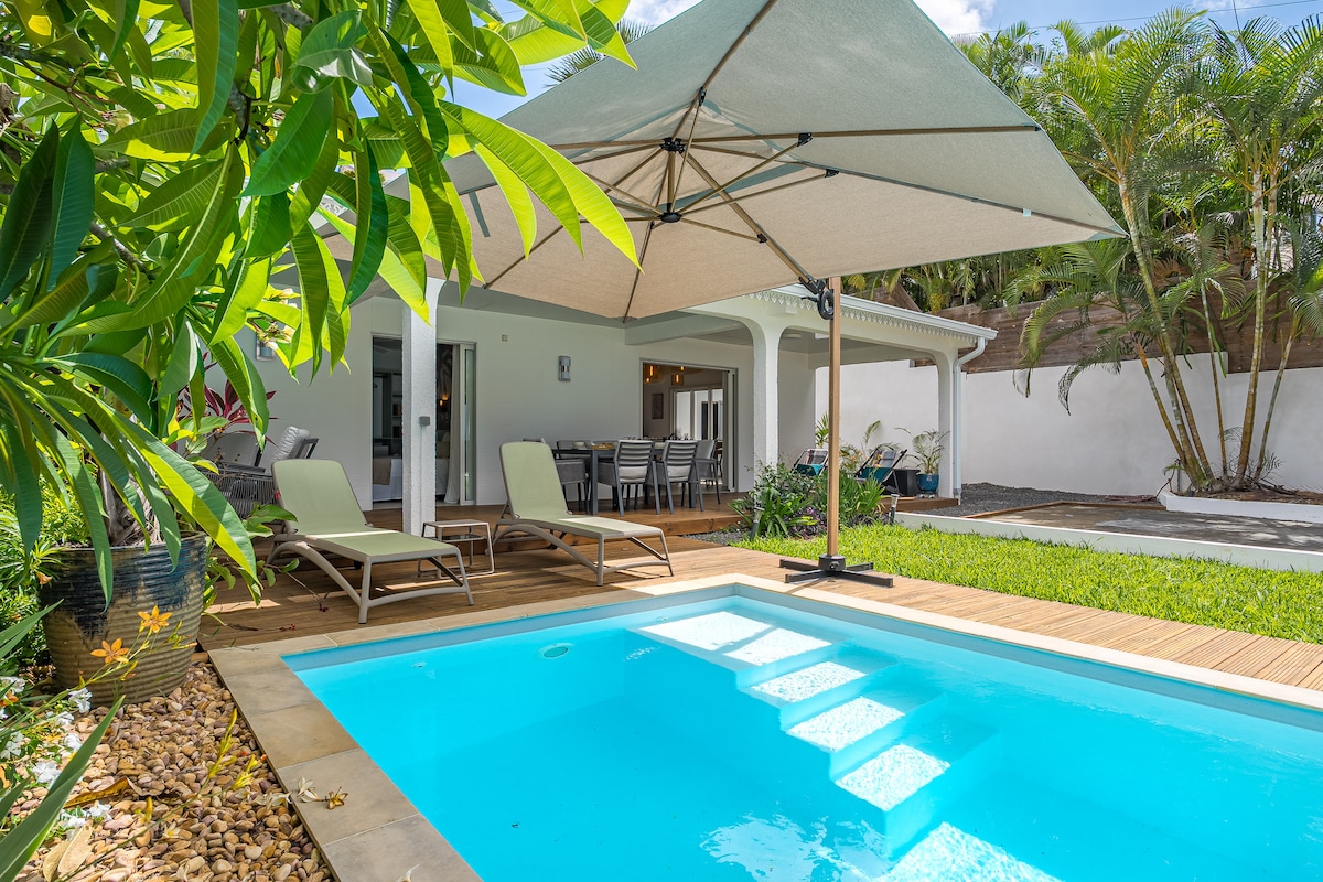 A tranquil outdoor area showcases a heated pool with inviting blue water, complemented by wooden decking and comfortable lounge chairs. An umbrella provides shade, while a dining table is visible in the background, surrounded by lush greenery and tropical plants.
