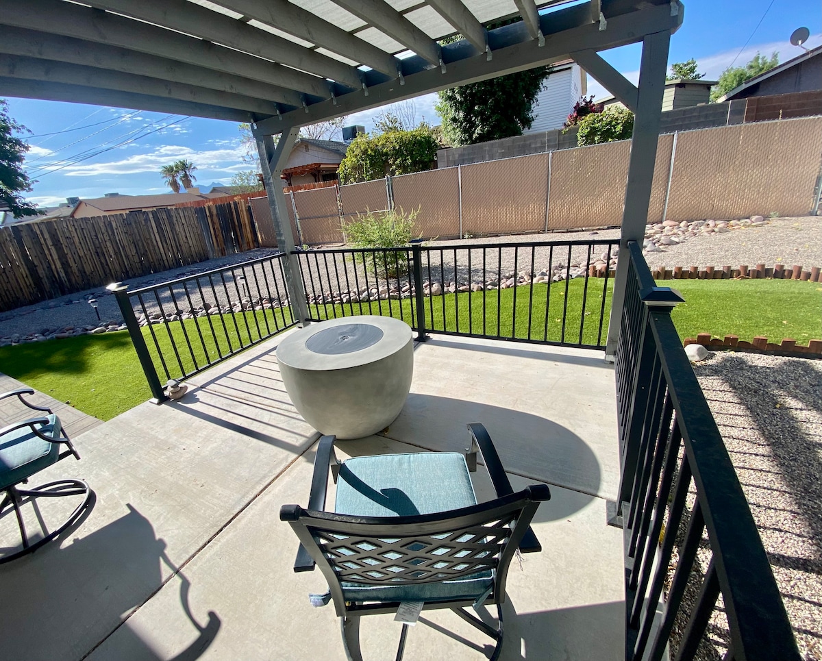 A shaded outdoor patio area features a round stone table accompanied by a single relaxed chair. The surrounding space is neatly landscaped with gravel, small plants, and low fencing, offering a private atmosphere. Tall fencing provides additional seclusion from adjacent properties.
