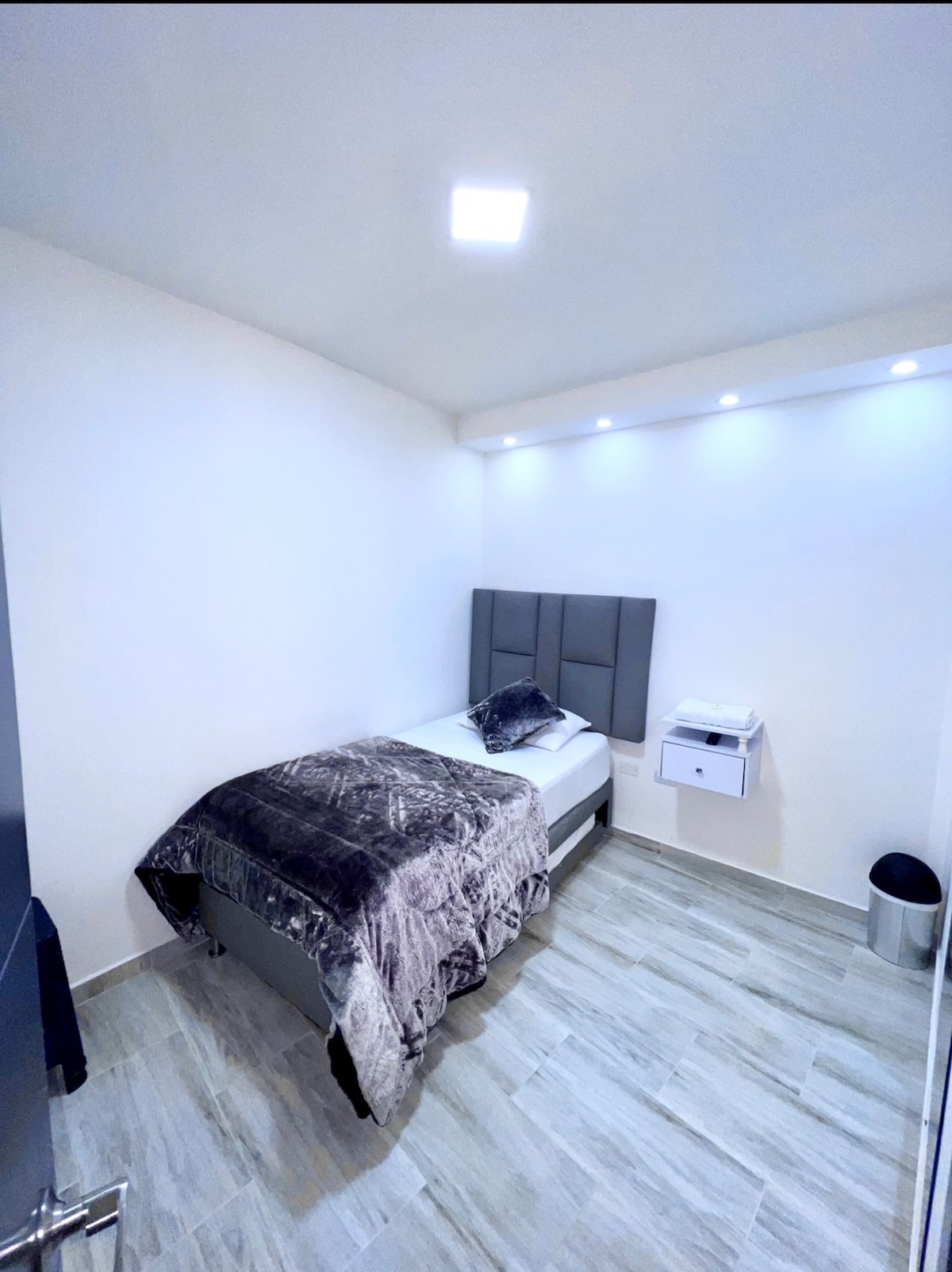 A compact sleeping area is highlighted by a single bed covered with a textured gray blanket. Soft lighting is provided by recessed fixtures above, illuminating the minimalist decor. A nightstand is mounted on the wall, while a wastebasket sits discreetly in the corner.