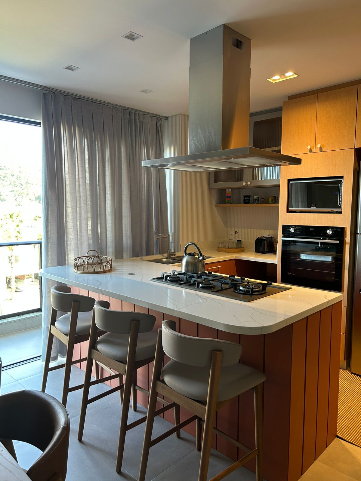 A modern kitchen features a central island with four stools, allowing for casual seating. A stovetop and hood are positioned above the island, complemented by built-in appliances. Natural light floods the space through a large window with sheer curtains, enhancing the inviting atmosphere.
