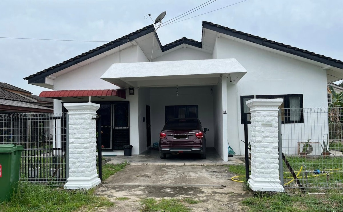 A modern house exterior features a white façade with a sloped roof. A covered porch leads to the entrance, and a parked car is visible in the driveway. The property is enclosed by a fence with stone pillars, and grassy areas surround the structure.