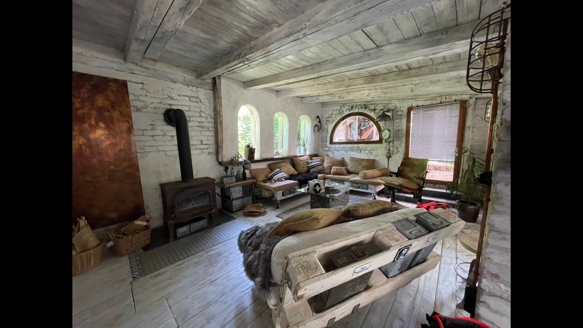 A spacious living area features a mix of comfortable seating options arranged around a central table. Natural light filters through arched windows, illuminating the rustic decor and wooden beams. A stove adds a cozy touch, while potted plants enhance the inviting atmosphere.