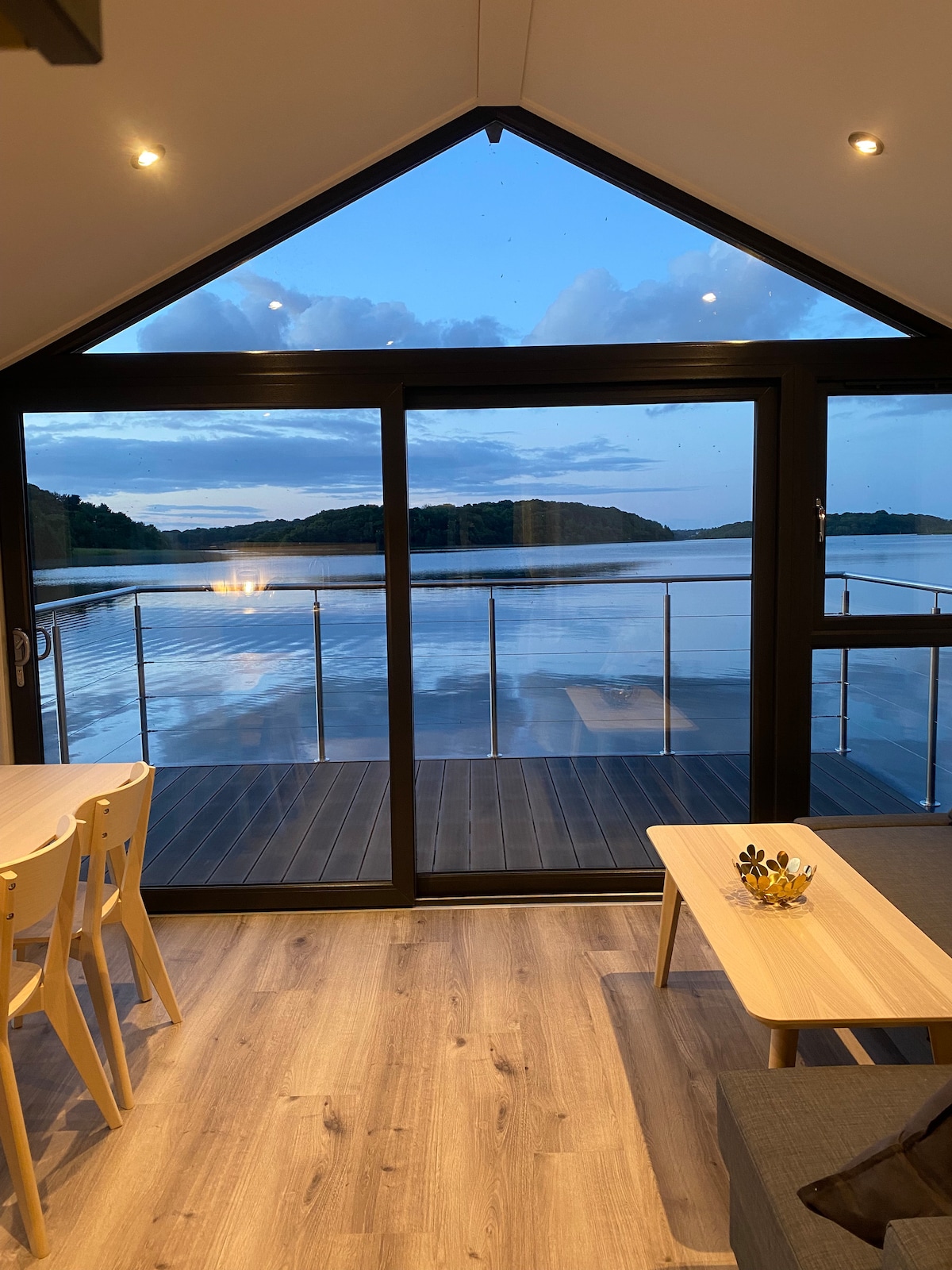 Large glass doors frame a view of the serene Lough Erne, with soft clouds reflected on the water's surface. The nearby dining area features a light wooden table paired with matching chairs, while a coffee table rests in the cozy living space.