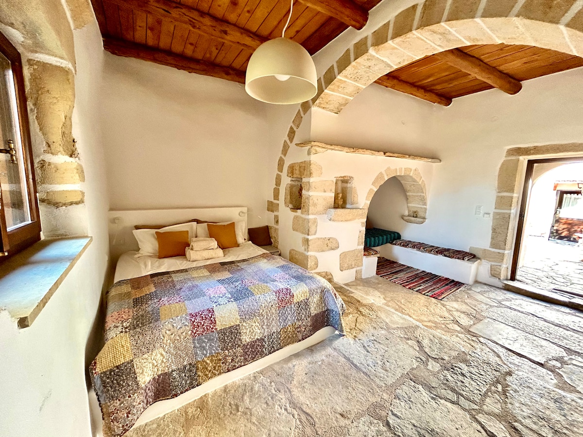 A cozy bedroom features a spacious bed adorned with a patchwork quilt. Natural light enters through a window, while a warm glow emanates from a rounded pendant light. An arched niche provides additional space for relaxation and a comfortable seating area is visible along the wall.