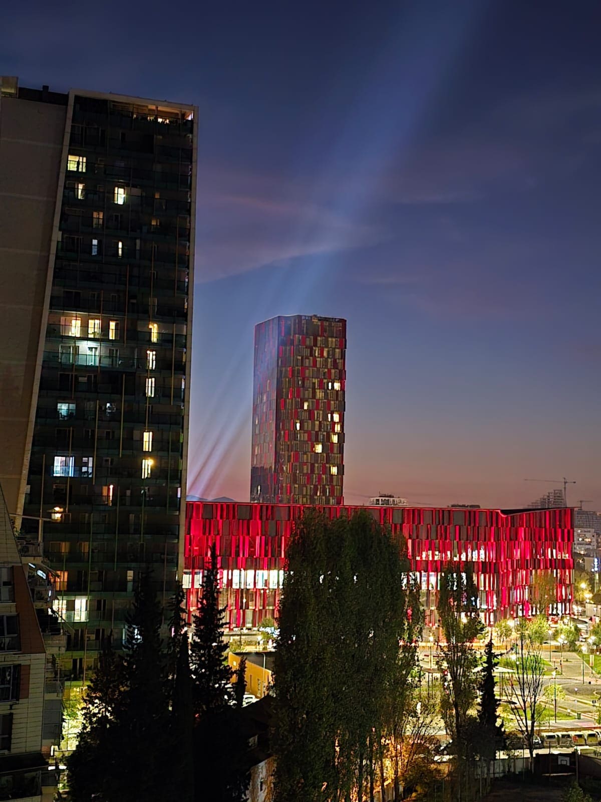 The image shows a modern urban landscape at dusk, featuring a striking tower illuminated with red lights. Surrounding buildings are partially visible, with trees in the foreground adding depth to the scene. A soft gradient in the sky transitions from twilight to night.