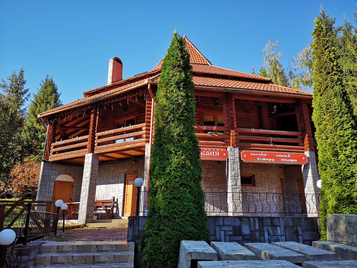 A multi-level chalet showcases a blend of wooden and stone architecture. The structure features a red roof and expansive patios framed by neatly trimmed evergreen trees. Soft lighting elements line the exterior, creating a welcoming entrance.